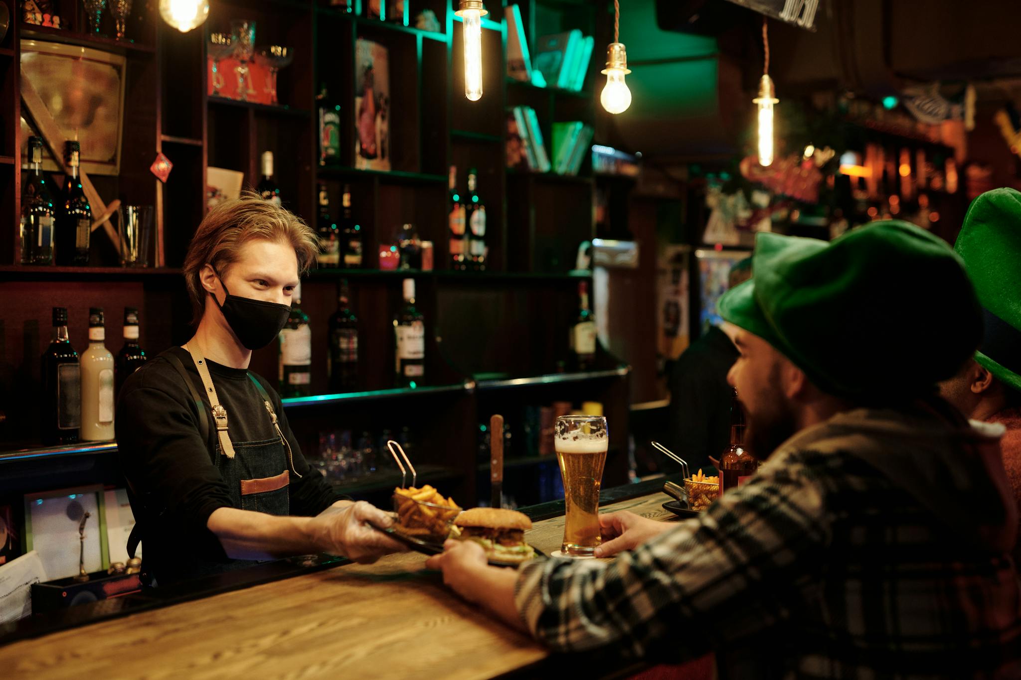 Masked bartender serves burger and beer at a lively pub during festive celebration.