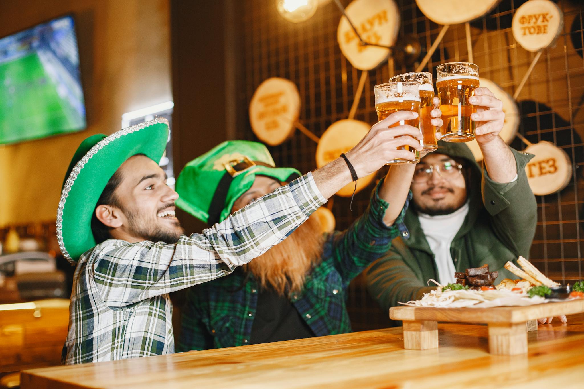 Three friends in Irish attire toasting with beers at a lively pub festival.