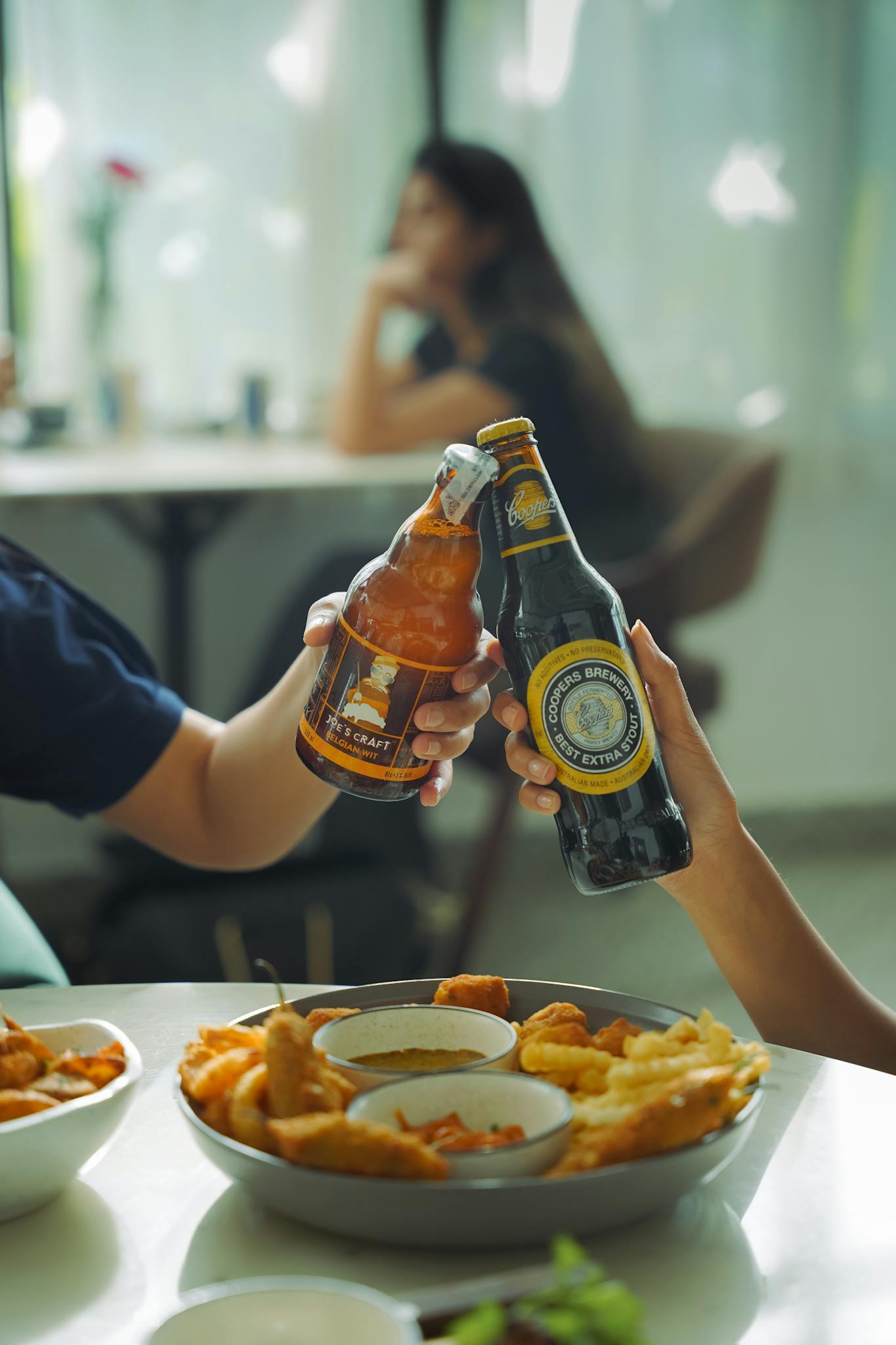 Two people toasting with craft beer bottles over a table of snacks.