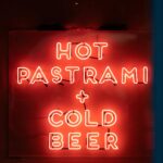 Vibrant neon sign in London offering hot pastrami and cold beer, creating a warm ambiance.